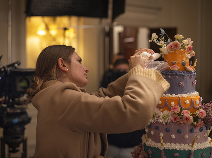A woman carefully decorates a tall, multi-tiered cake with flowers and icing using a piping bag, while a film camera and lighting equipment are set up nearby, indicating a behind-the-scenes production setting.
