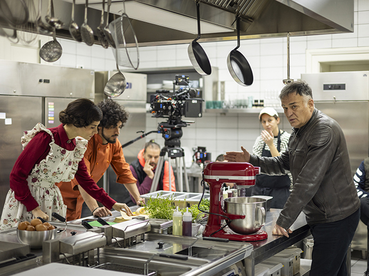 A film crew shoots a scene in a commercial kitchen, where actors and crew members gather around a stainless-steel counter with cooking equipment, including a red stand mixer, while a man gestures as if giving direction and a camera films the action in the background.