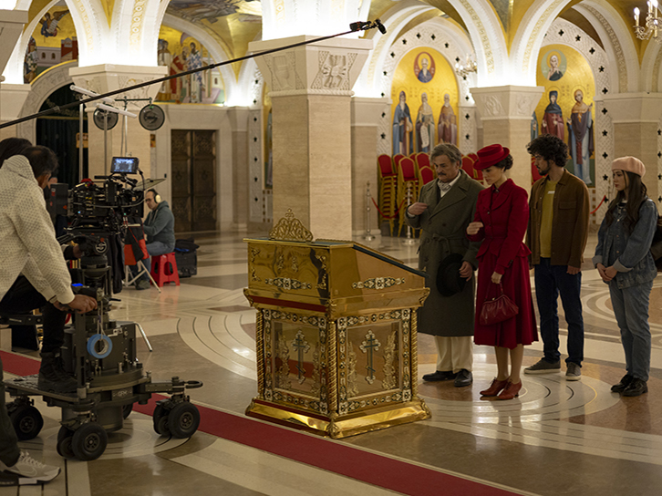 A film crew records a scene inside an ornate church, where several actors in period-style clothing stand solemnly beside a decorative golden casket, while a camera operator and boom microphone capture the moment.